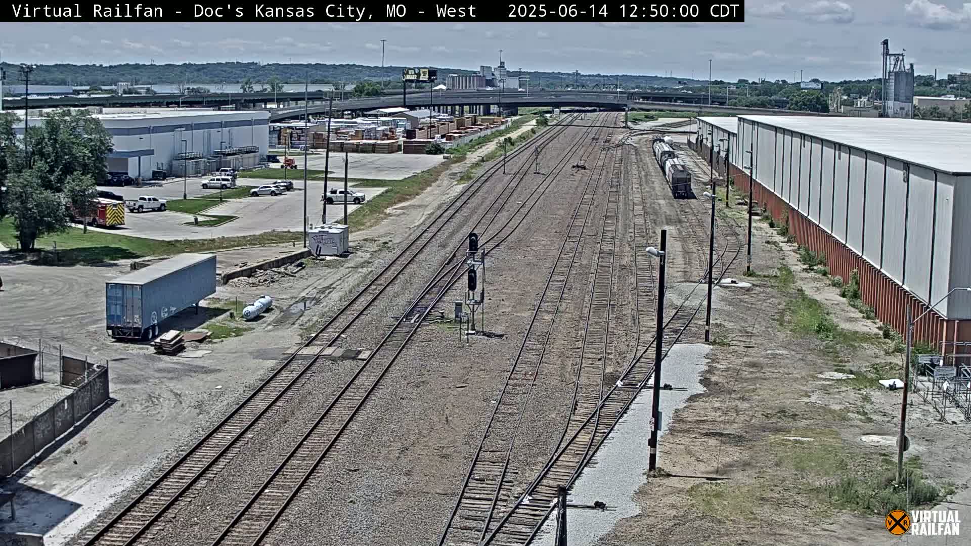 A high-angle view of a railway yard on a partly sunny day, showing multiple train tracks, industrial buildings, and parked vehicles.