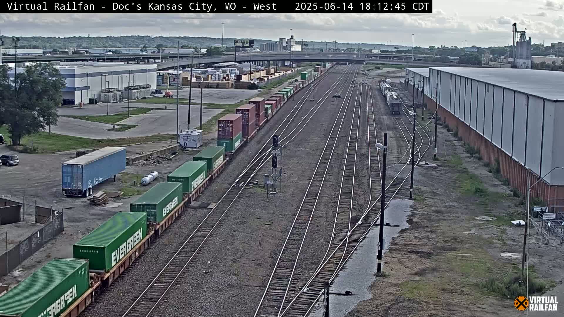 A high-angle view of a railway yard on a partly sunny day, showing multiple train tracks, industrial buildings, and parked vehicles.