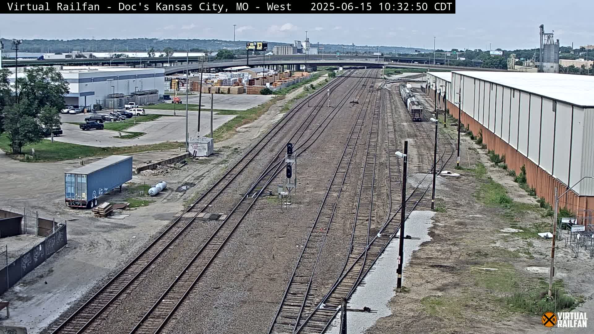 A high-angle view of a railway yard on a partly cloudy day, showing multiple train tracks, industrial buildings, and a lone freight train.