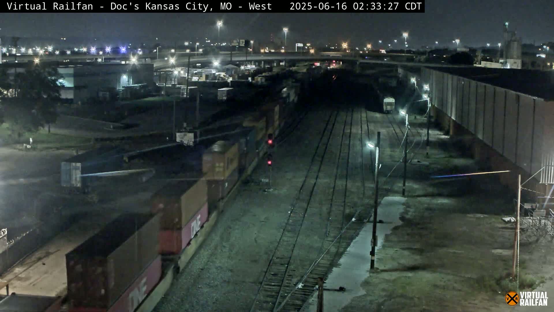 A nighttime, aerial view of a train yard with a long freight train alongside several railway tracks, under artificial light.
