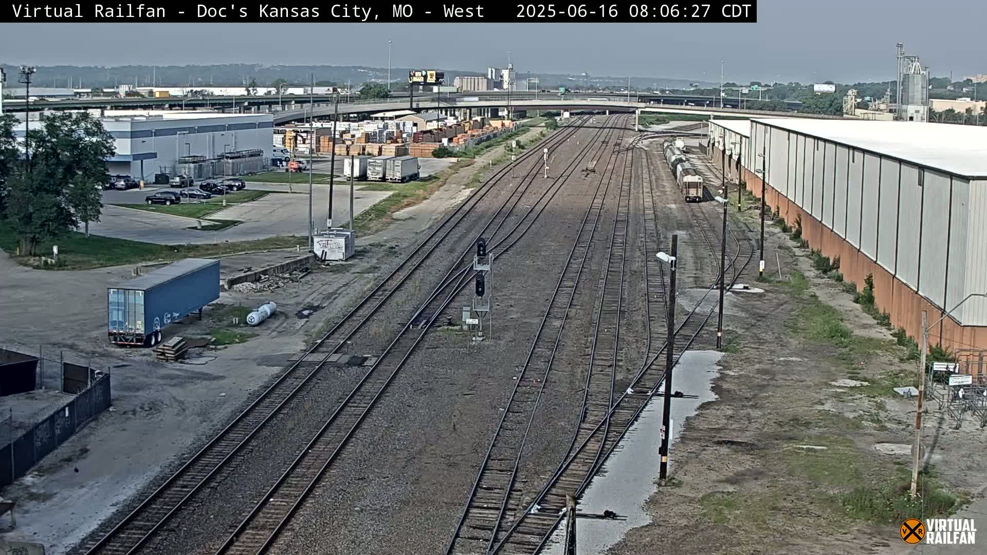 A high-angle view of railway tracks splitting into several directions, surrounded by industrial buildings and parked vehicles on a partly sunny day.
