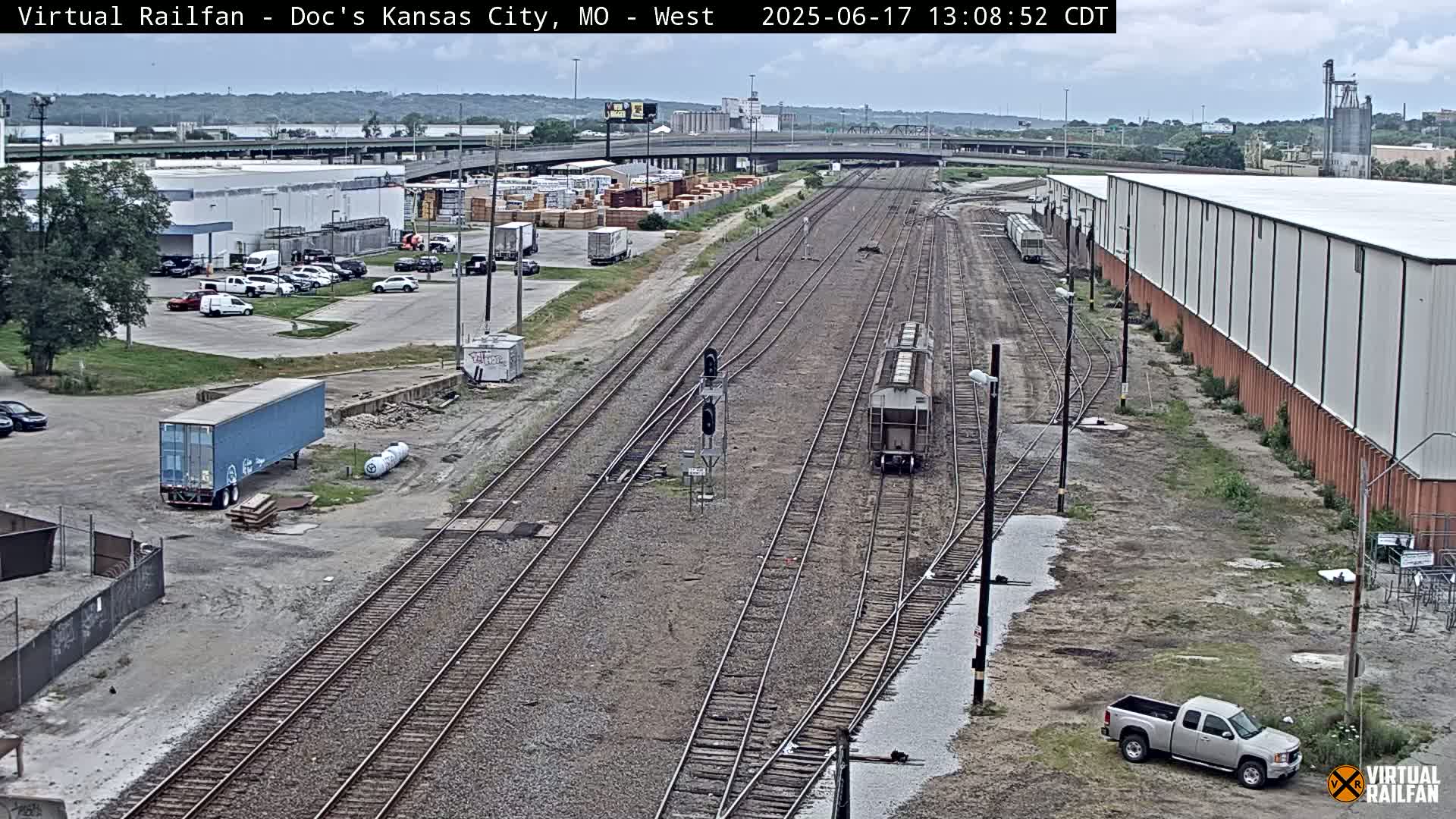A view of railroad tracks with a freight car and several buildings and vehicles in a parking lot on an overcast day.