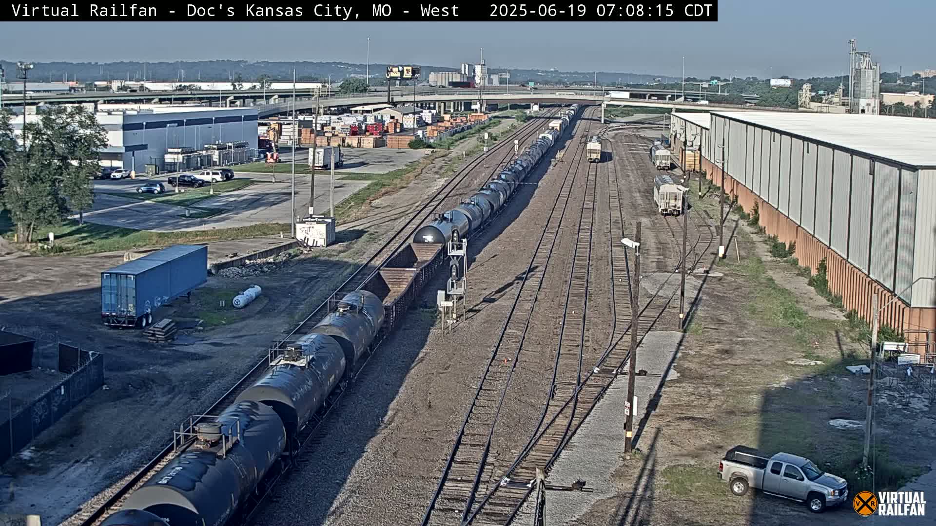 A long freight train of tanker cars and boxcars travels along multiple railway tracks on a sunny day near industrial buildings and a highway overpass.