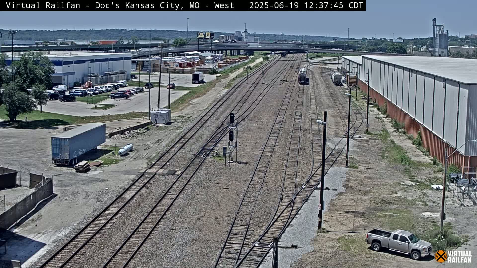 A high-angle view of a railway track on a sunny day, with industrial buildings, parked vehicles, and a gray shipping container visible in the surrounding area.