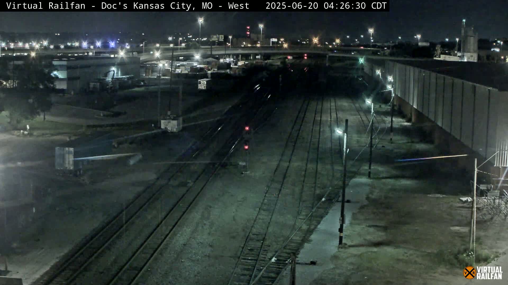 A nighttime view of railroad tracks, industrial buildings, and a highway overpass under clear skies.