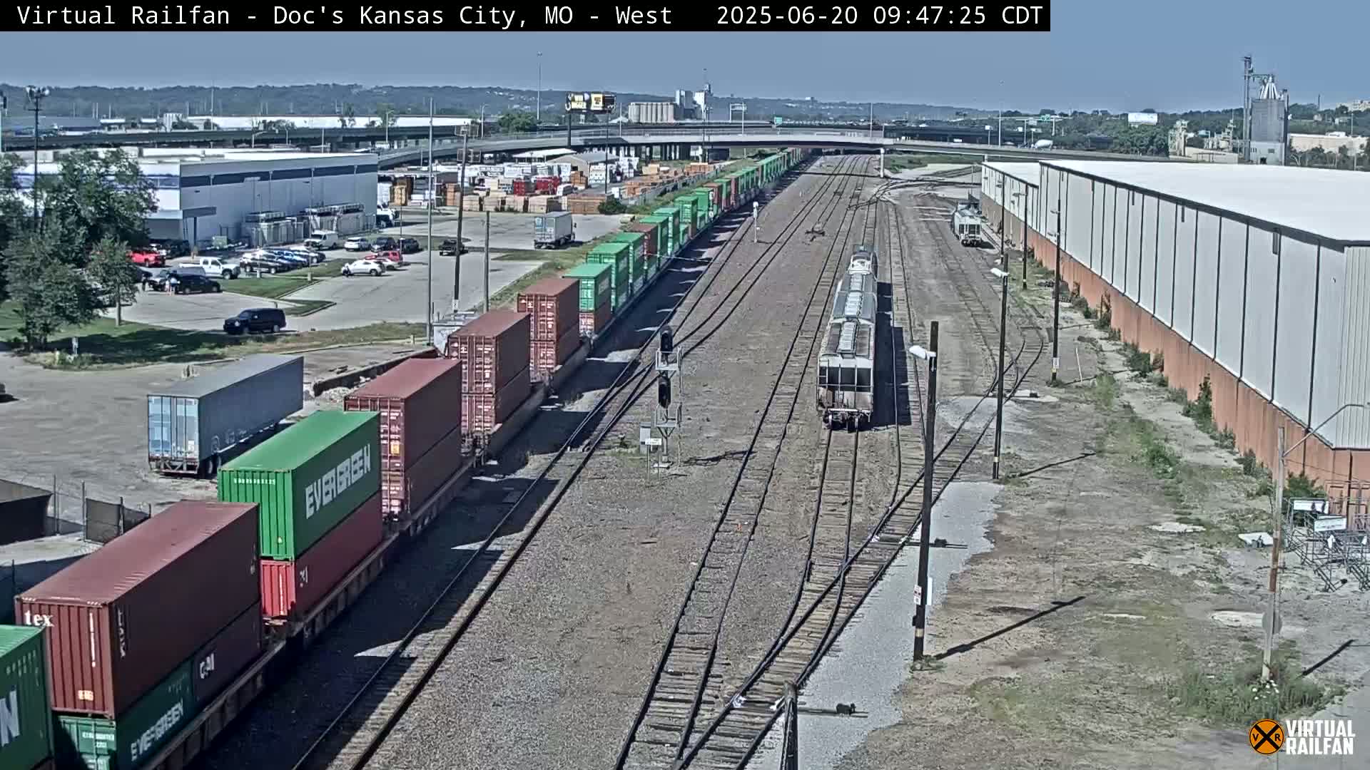 A long freight train carrying numerous shipping containers is seen on a railway track next to industrial buildings and a parking lot under a mostly sunny sky.