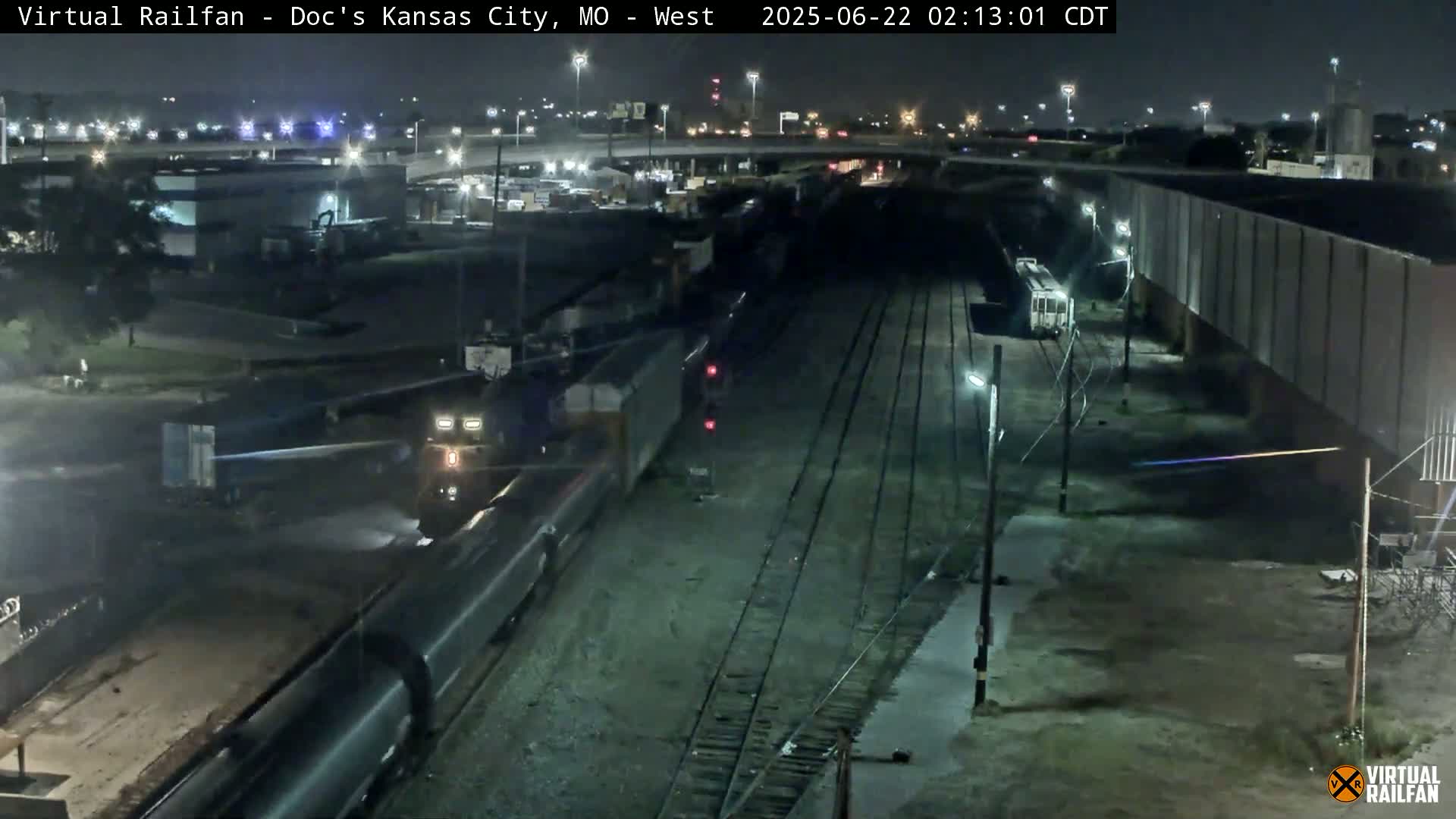 A nighttime view of a freight train moving through a rail yard, under clear skies.