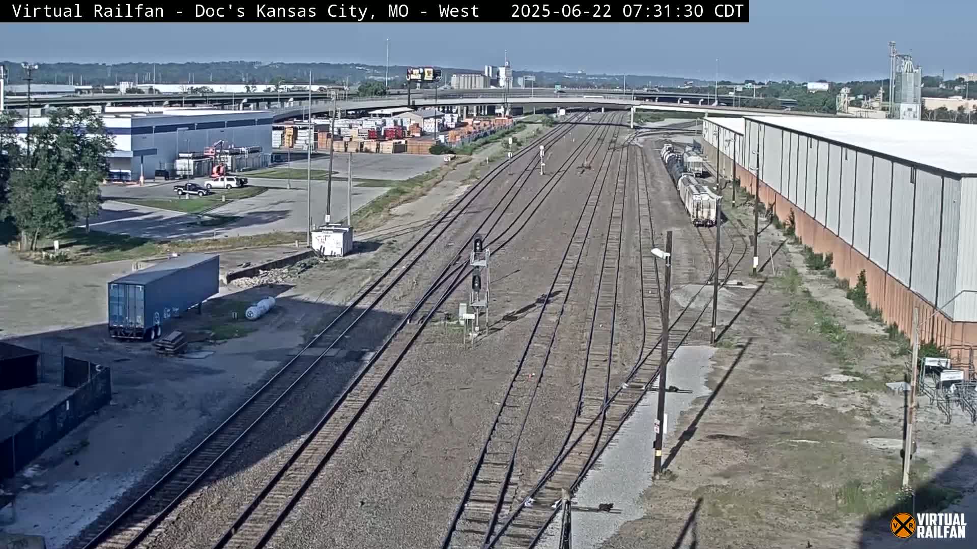 A sunny view of railroad tracks and industrial buildings, with several rail cars parked nearby.