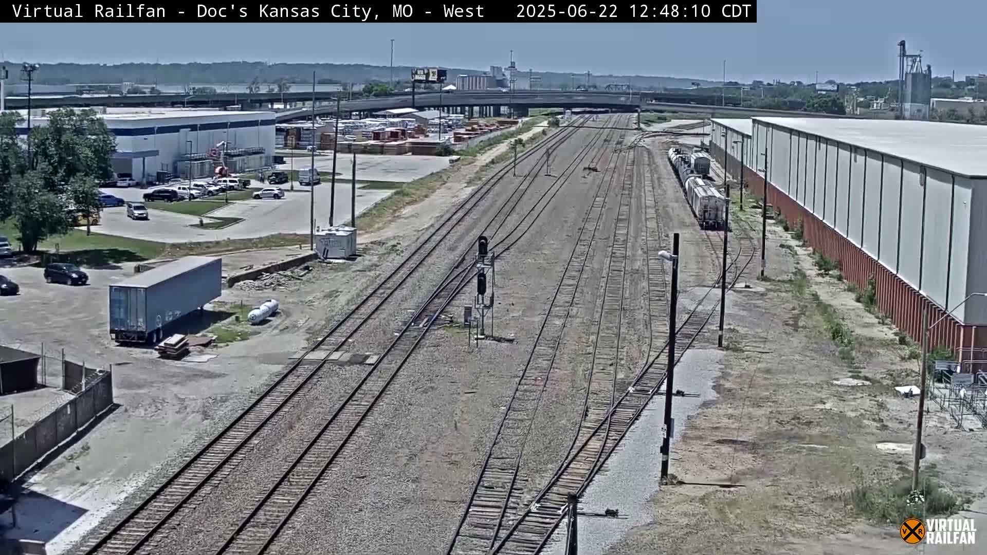 A long freight train is visible on a railway track next to industrial buildings and parking lots on a sunny day.