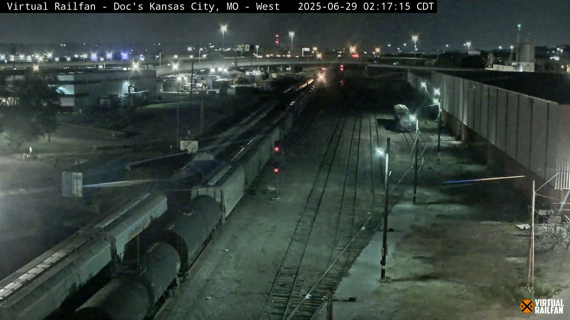 A long freight train sits on railway tracks at night near a highway overpass.