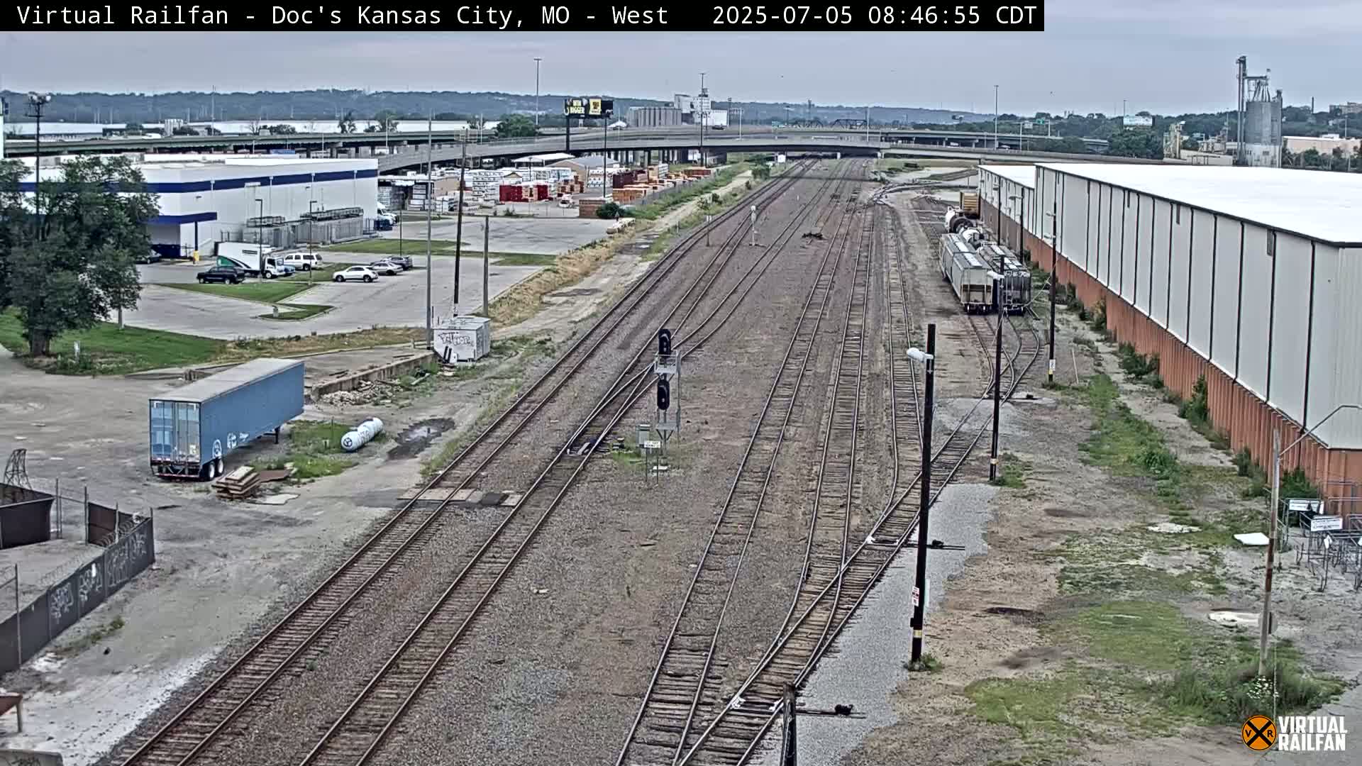 A view of railroad tracks extending from a parking lot with several buildings and a train car, under an overcast sky.