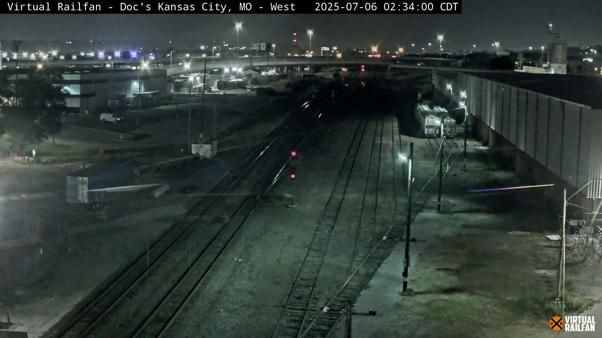 A nighttime view of railroad tracks and industrial buildings, with a train visible in the distance under a clear sky.