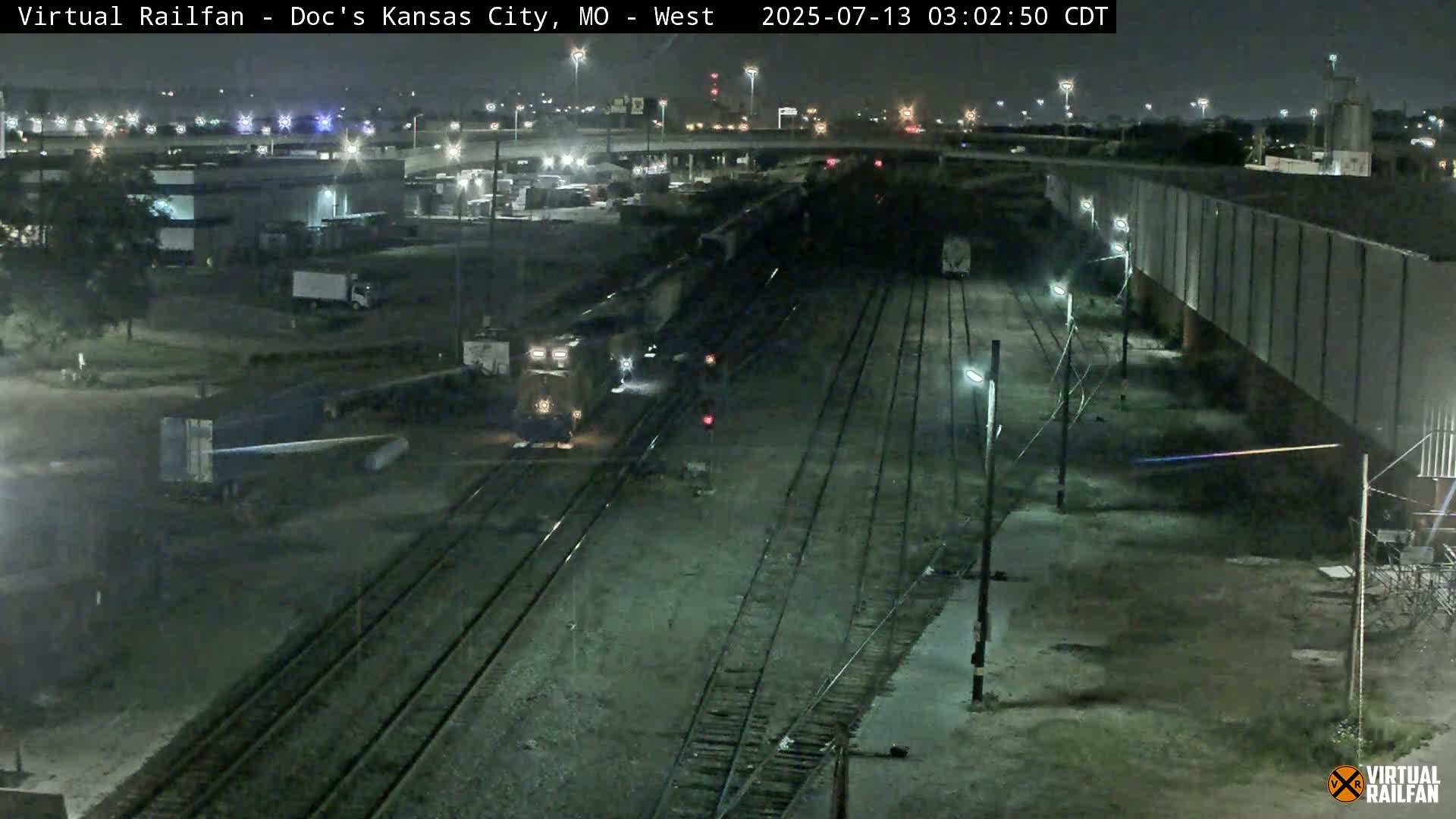 A nighttime view of a train passing through a rail yard, with several other railcars visible nearby.