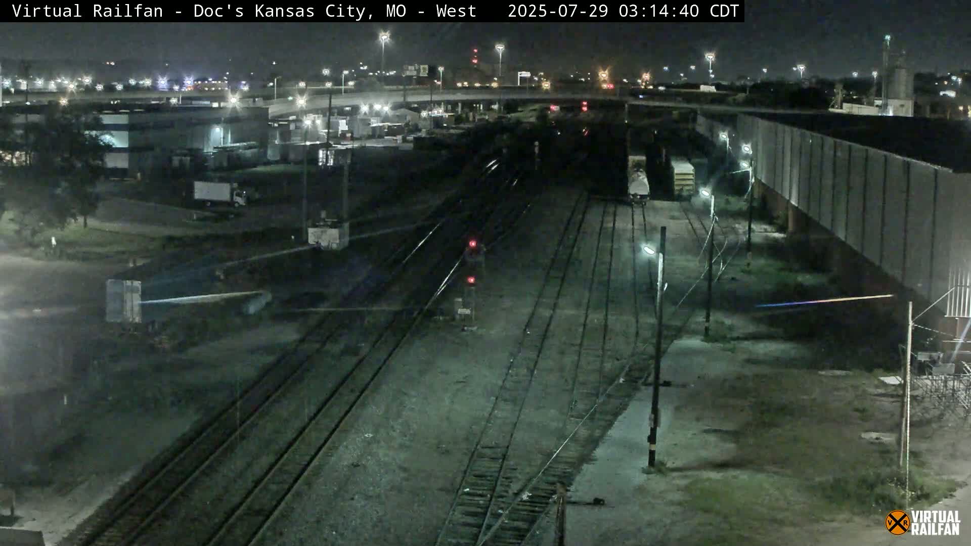 A nighttime view of a train yard with multiple train tracks, some freight cars, and industrial buildings under a clear sky.
