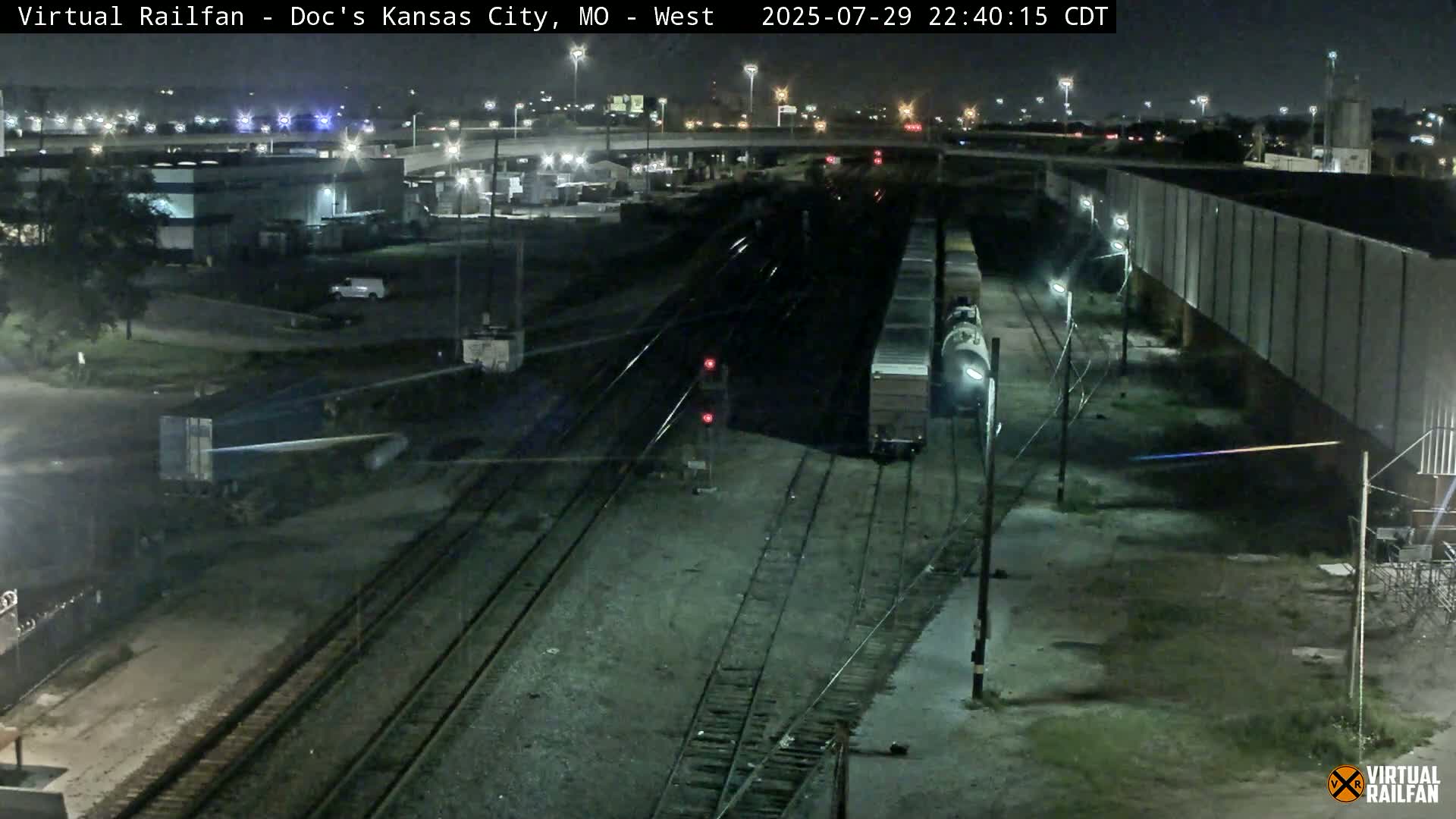 A nighttime view of a train yard with multiple train tracks, some freight cars, and industrial buildings under a clear sky.