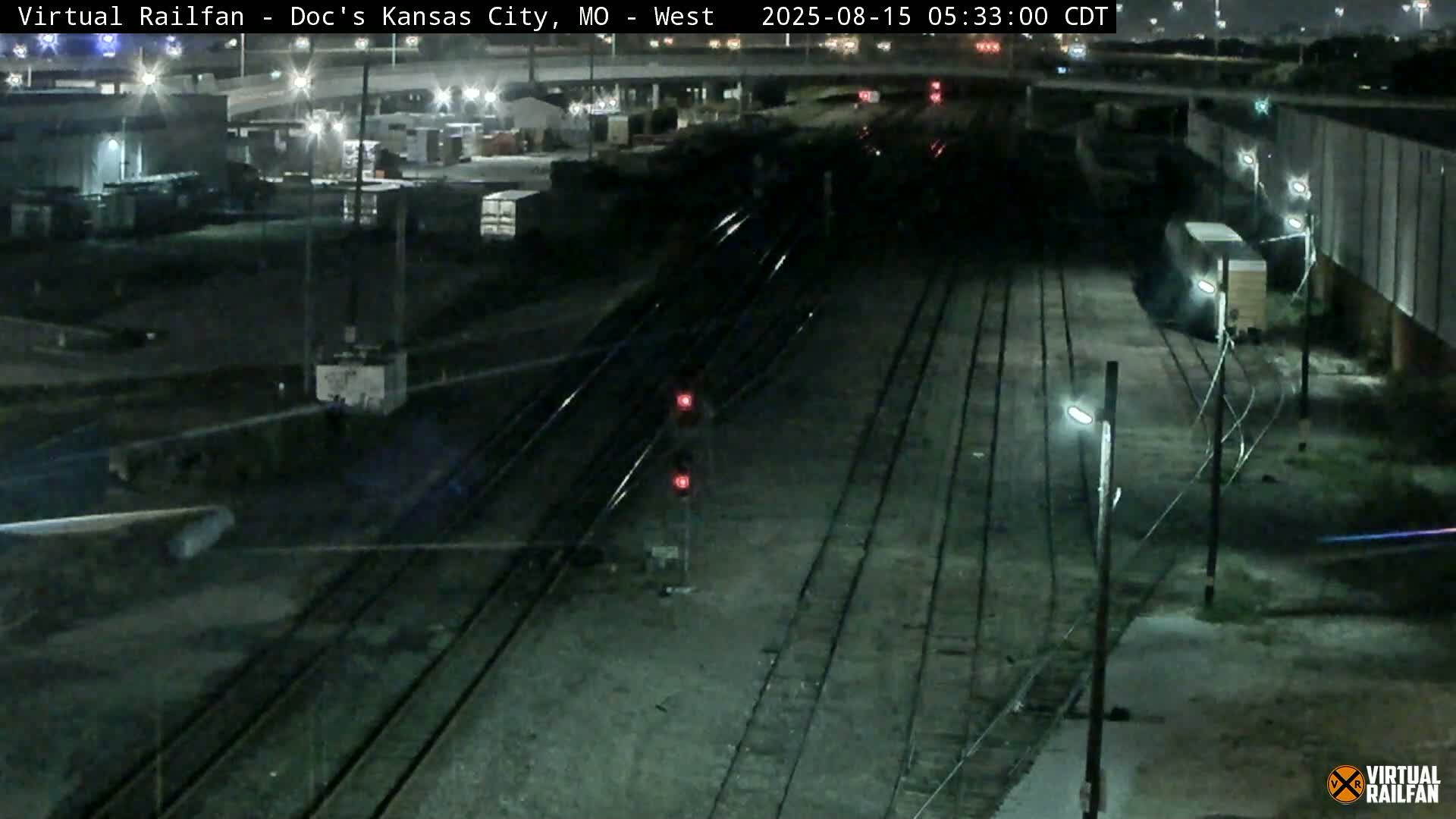 A nighttime view of a train yard with multiple train tracks, some freight cars, and industrial buildings under a clear sky.