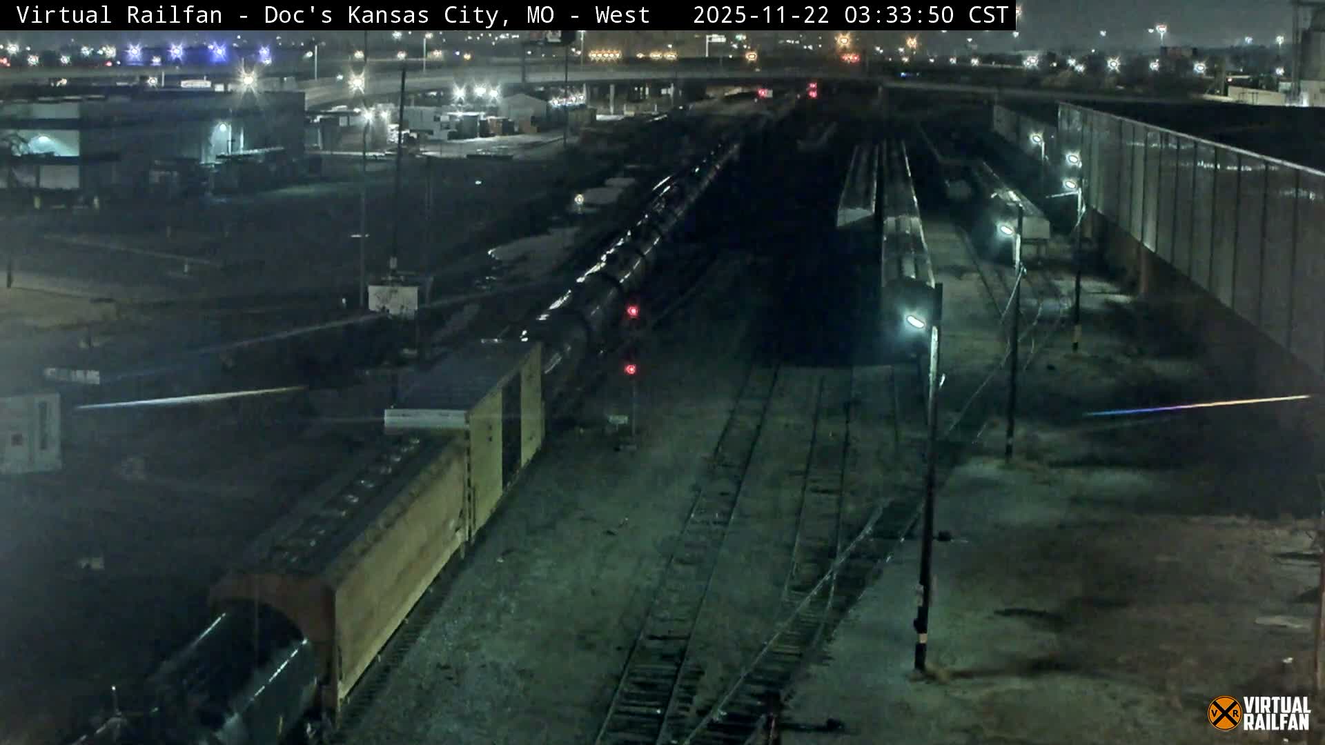 An elevated view reveals a bustling train yard at night with several trains on multiple tracks, illuminated by bright artificial lights under clear conditions, alongside industrial buildings and distant city lights.