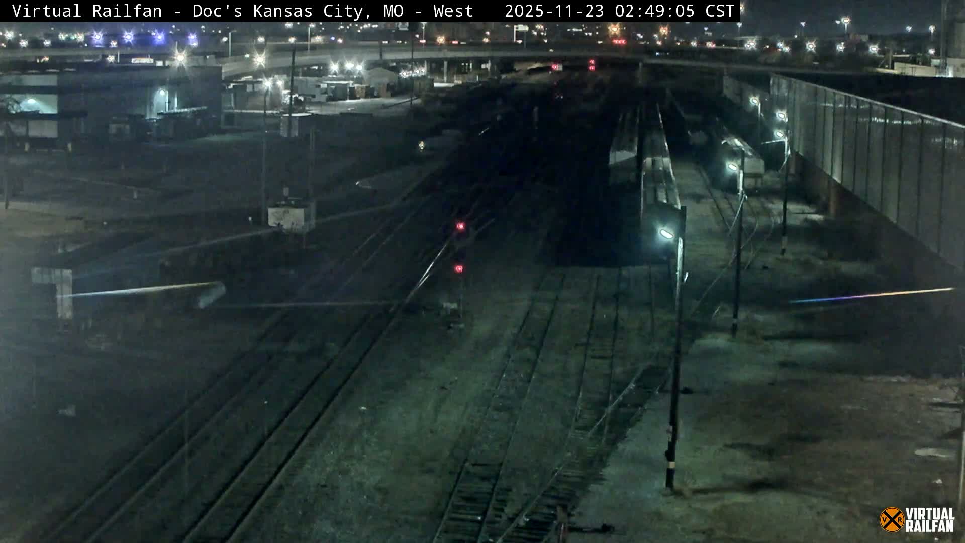 A wide, nighttime view of an extensive railway yard under clear skies, featuring numerous tracks, red signal lights, bright utility poles, and distant city lights and structures, with faint rail cars visible on the right.