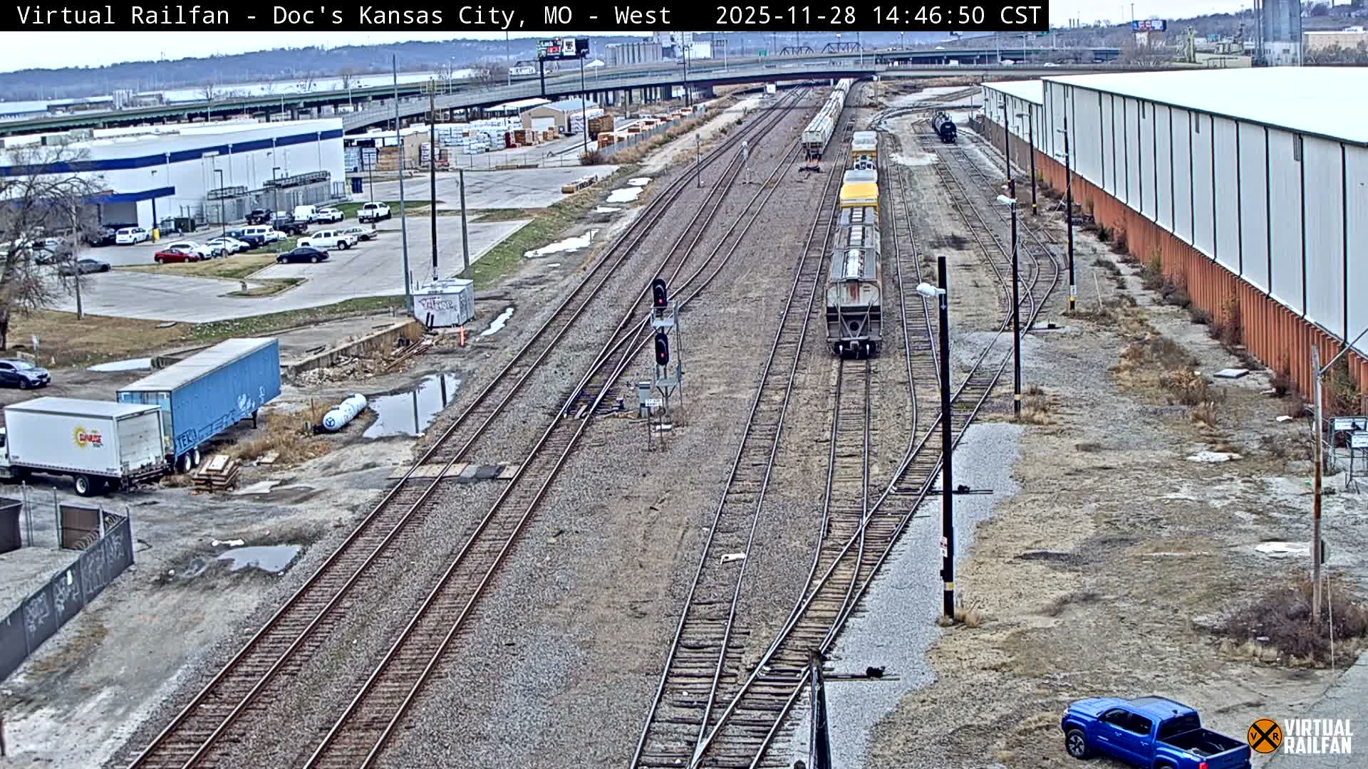 A wide overhead view captures an industrial railyard with numerous parallel train tracks, several stationary railcars, a long white and orange warehouse, and various vehicles on wet ground under an overcast sky.
