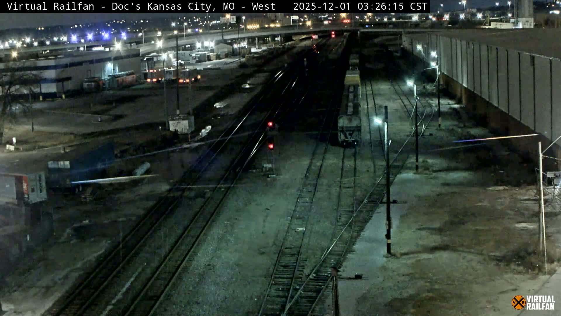 An elevated nighttime view presents a sprawling rail yard with multiple tracks, a parked train car, and two red signal lights, all brightly illuminated by industrial lighting and backdropped by distant city lights and a clear, dark sky.