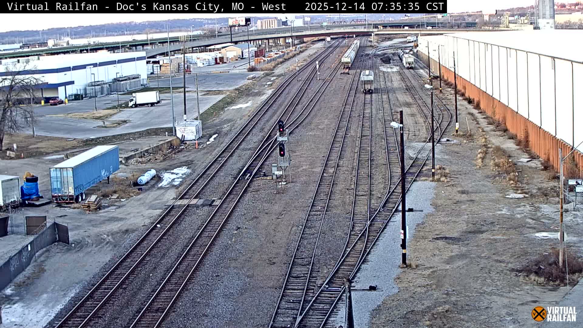 An elevated nighttime view presents a sprawling rail yard with multiple tracks, a parked train car, and two red signal lights, all brightly illuminated by industrial lighting and backdropped by distant city lights and a clear, dark sky.