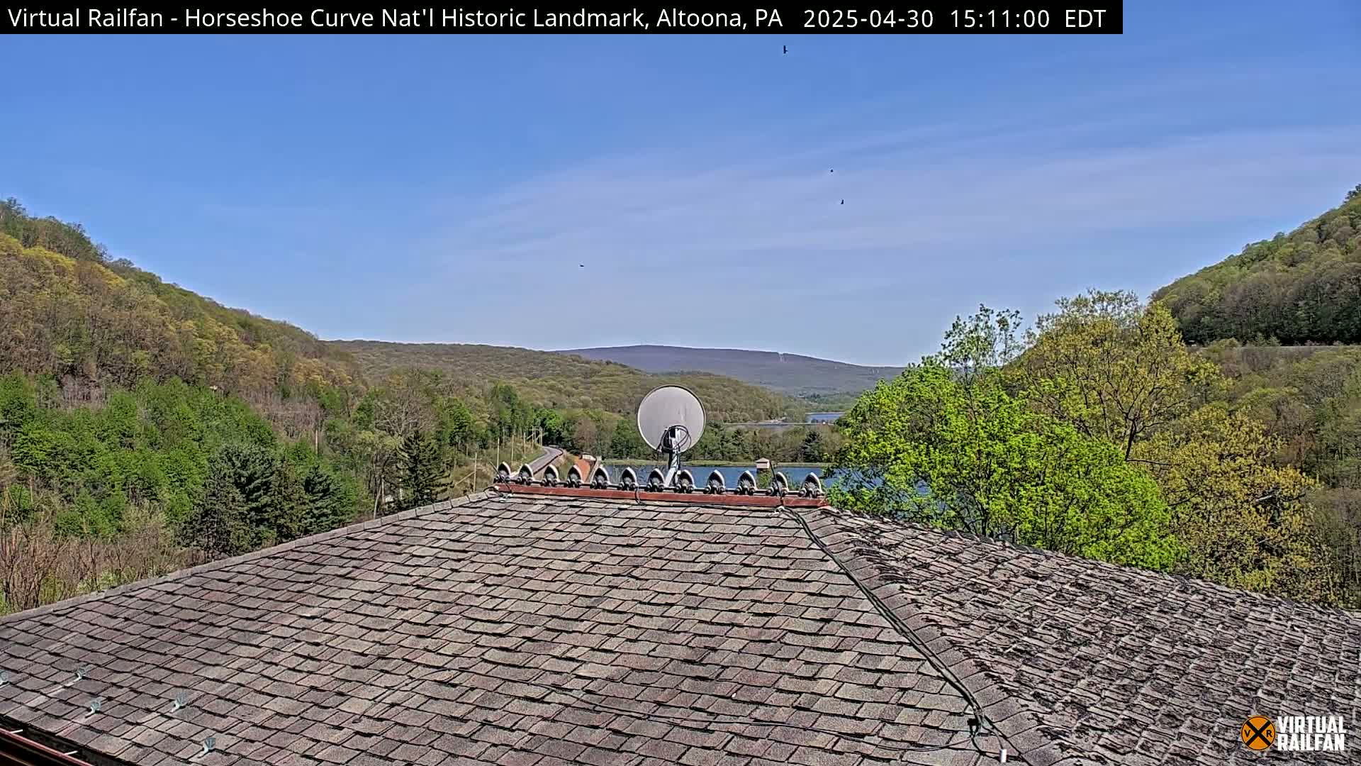 A satellite dish sits atop a shingled roof overlooking a valley of green trees and a body of water under a clear blue sky.