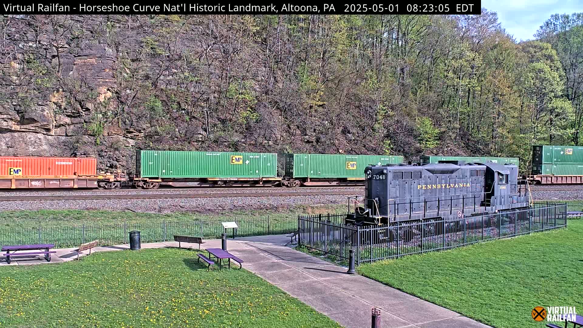 A freight train with green and orange shipping containers passes a dark-colored locomotive displayed behind a fence in a grassy park-like area on a sunny day.