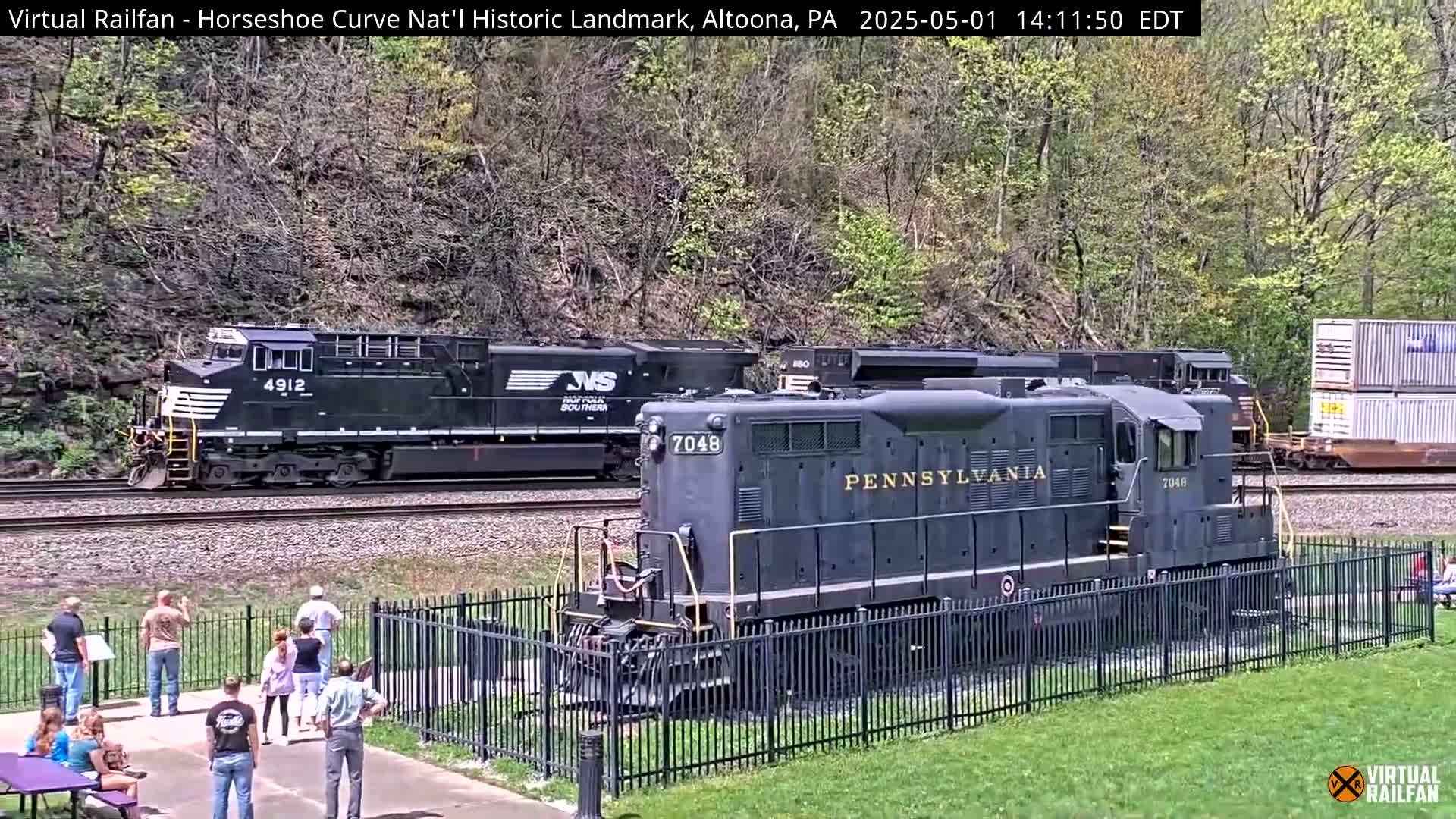 Two locomotives, one modern and one vintage, are seen on train tracks near a group of onlookers behind a black metal fence on a partly sunny day.