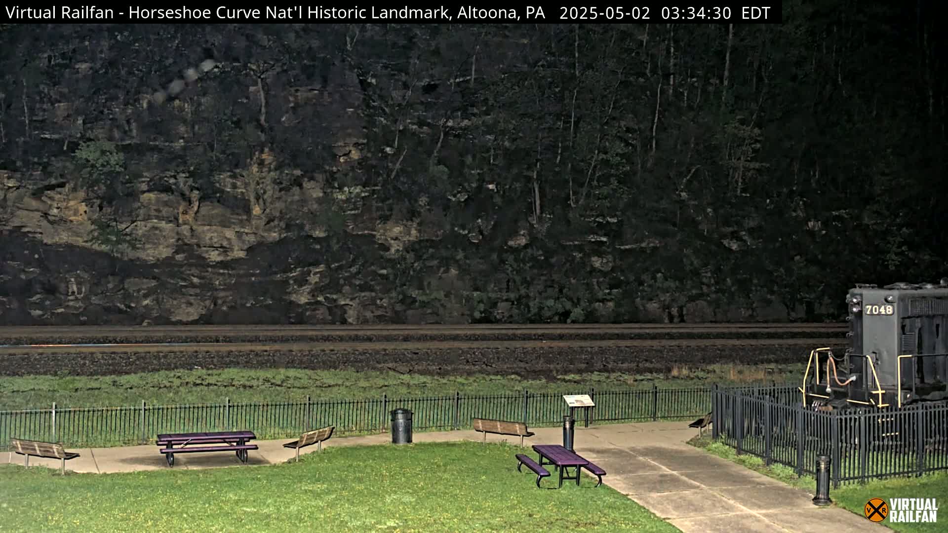 A nighttime view of a train partially visible behind a fence, next to a grassy area with picnic tables and benches, with a rocky hillside in the background.