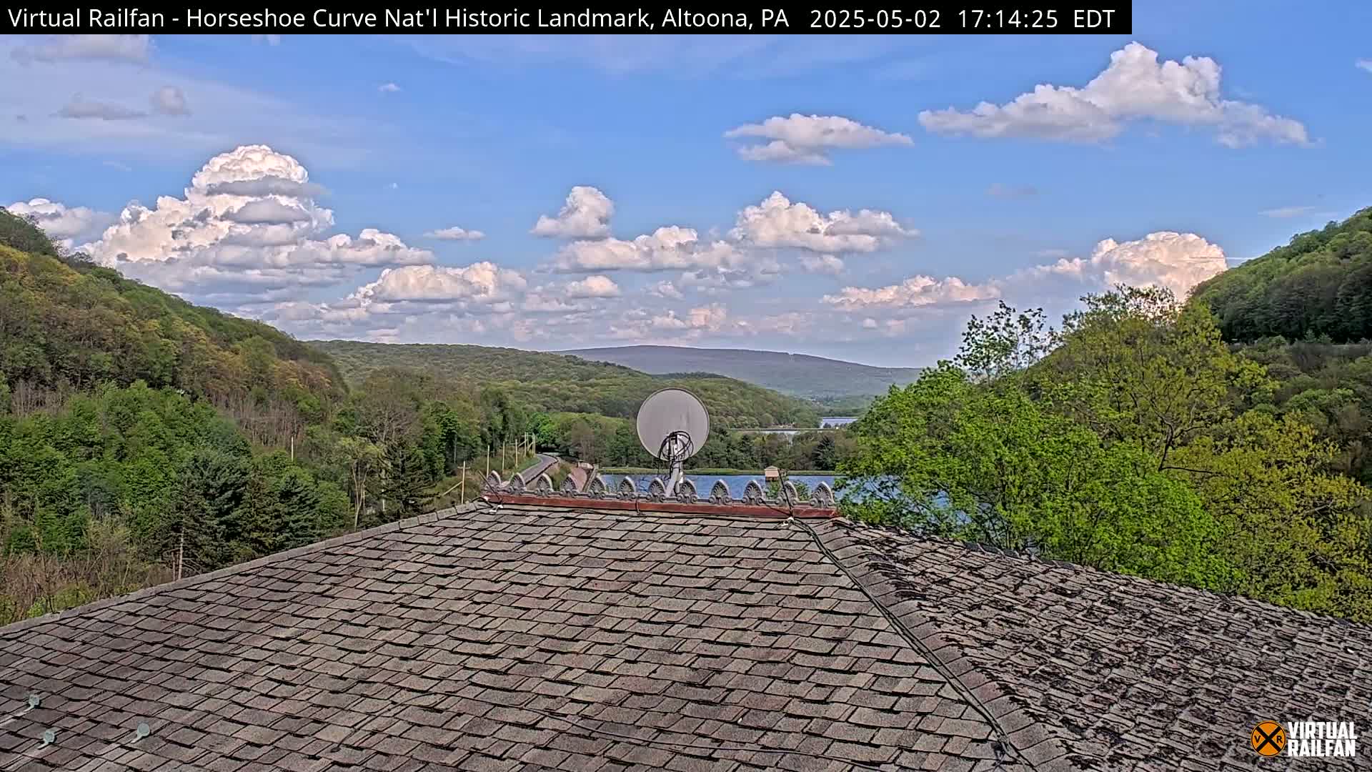 A partly cloudy sky overlooks a tree-covered hillside and a body of water, viewed from a weathered shingled roof with a satellite dish.