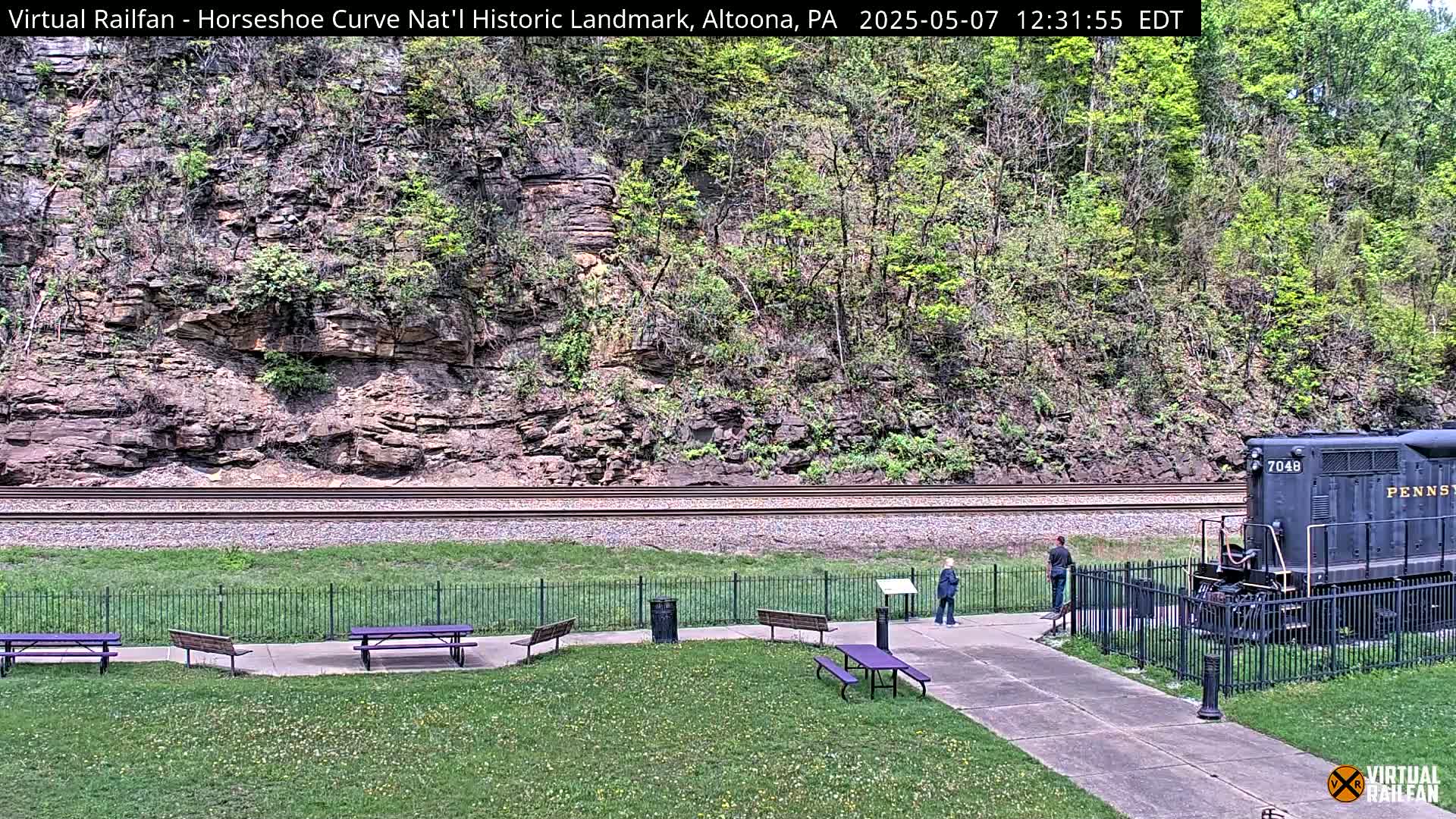 A sunny view of a grassy area with picnic tables, a black locomotive, and a rocky hillside beyond railroad tracks.