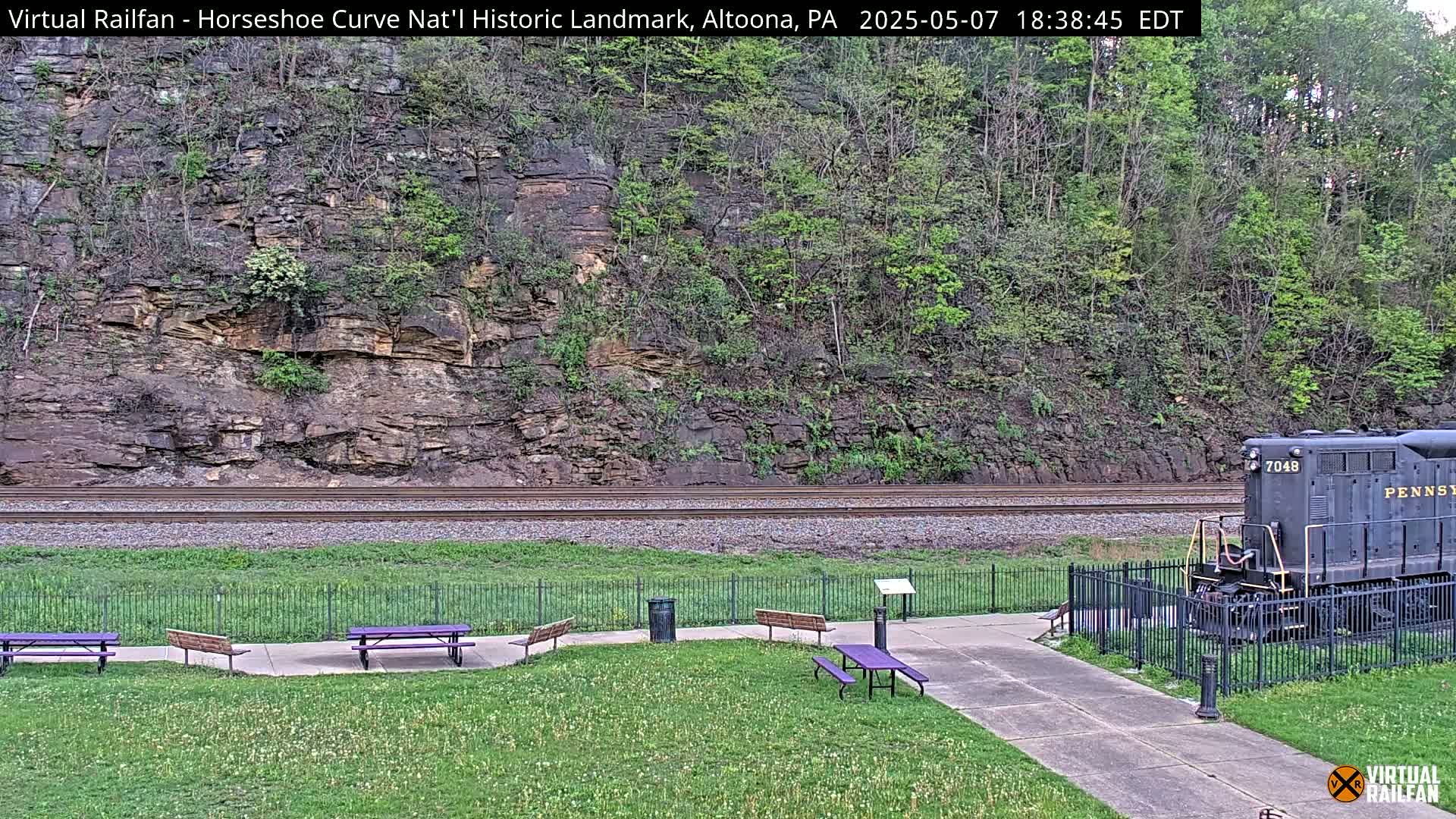 A dark-colored locomotive sits near a grassy area with picnic tables, next to railroad tracks that run alongside a rocky hillside covered in green trees and shrubs, under a daytime sky.