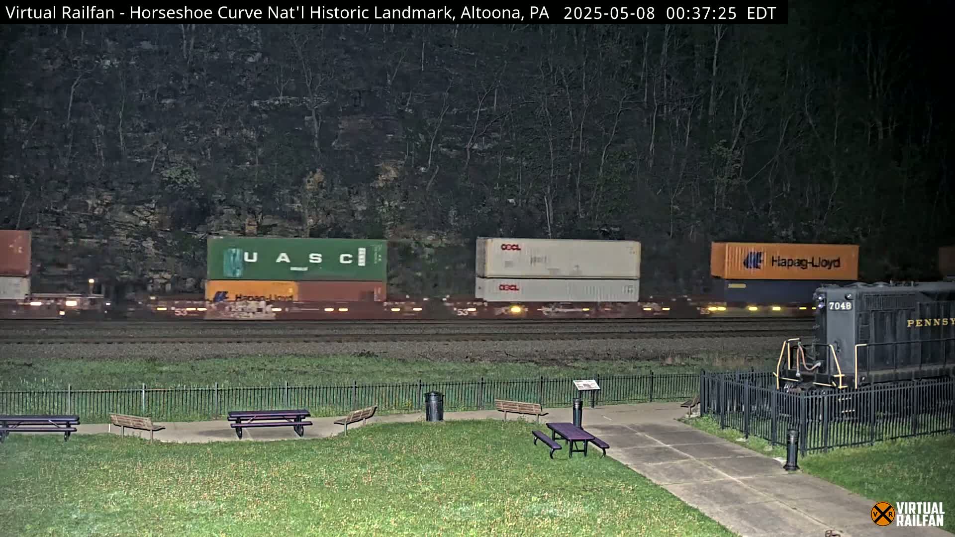 At night, a freight train with various shipping containers passes by a park with picnic tables and a black diesel locomotive.