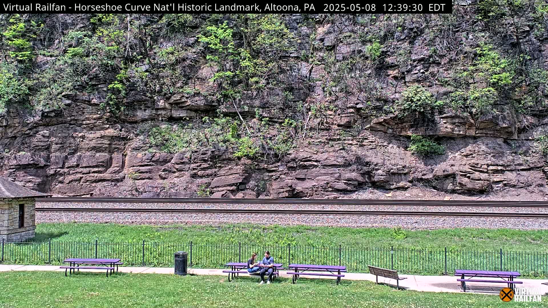 A rocky hillside with sparse vegetation overlooks a set of train tracks and a grassy area with picnic tables where two people are seated.