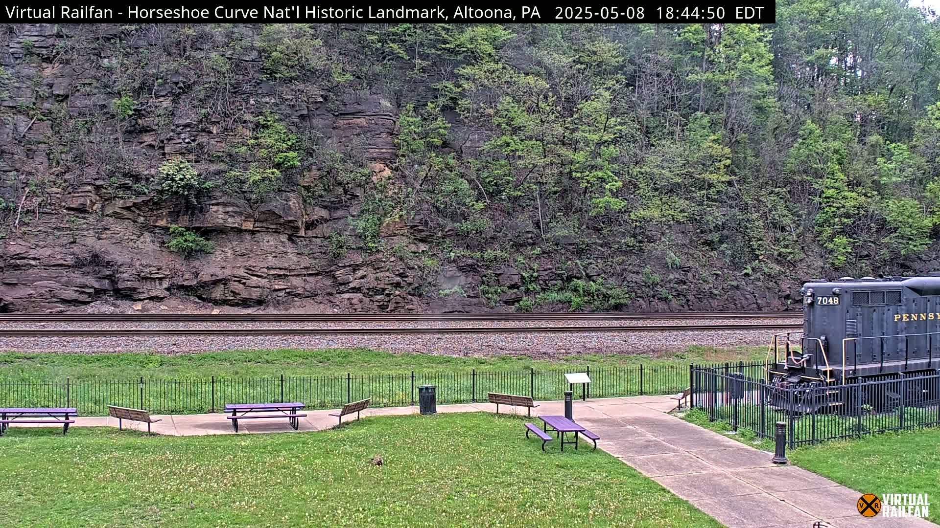 A dark-colored locomotive sits on railroad tracks beside a grassy area with picnic tables, a low metal fence, and a rocky hillside covered in green vegetation under an overcast sky.
