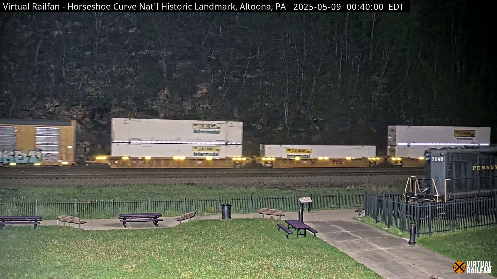 A nighttime view of a freight train passing by a grassy area with picnic tables, enclosed by a black metal fence, against a backdrop of dark trees.