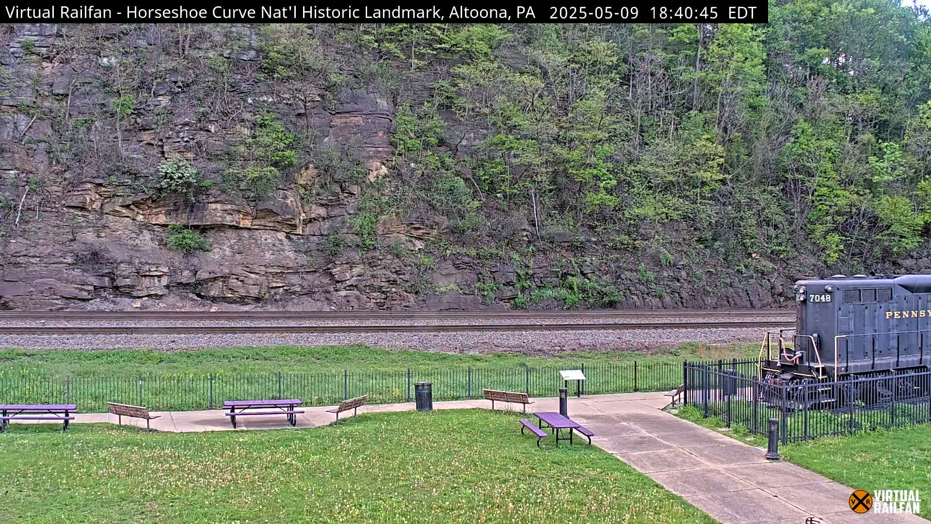 A grassy area with picnic tables and a paved path overlooks railroad tracks and a steep, rocky hillside covered in trees and underbrush, under a daytime sky.