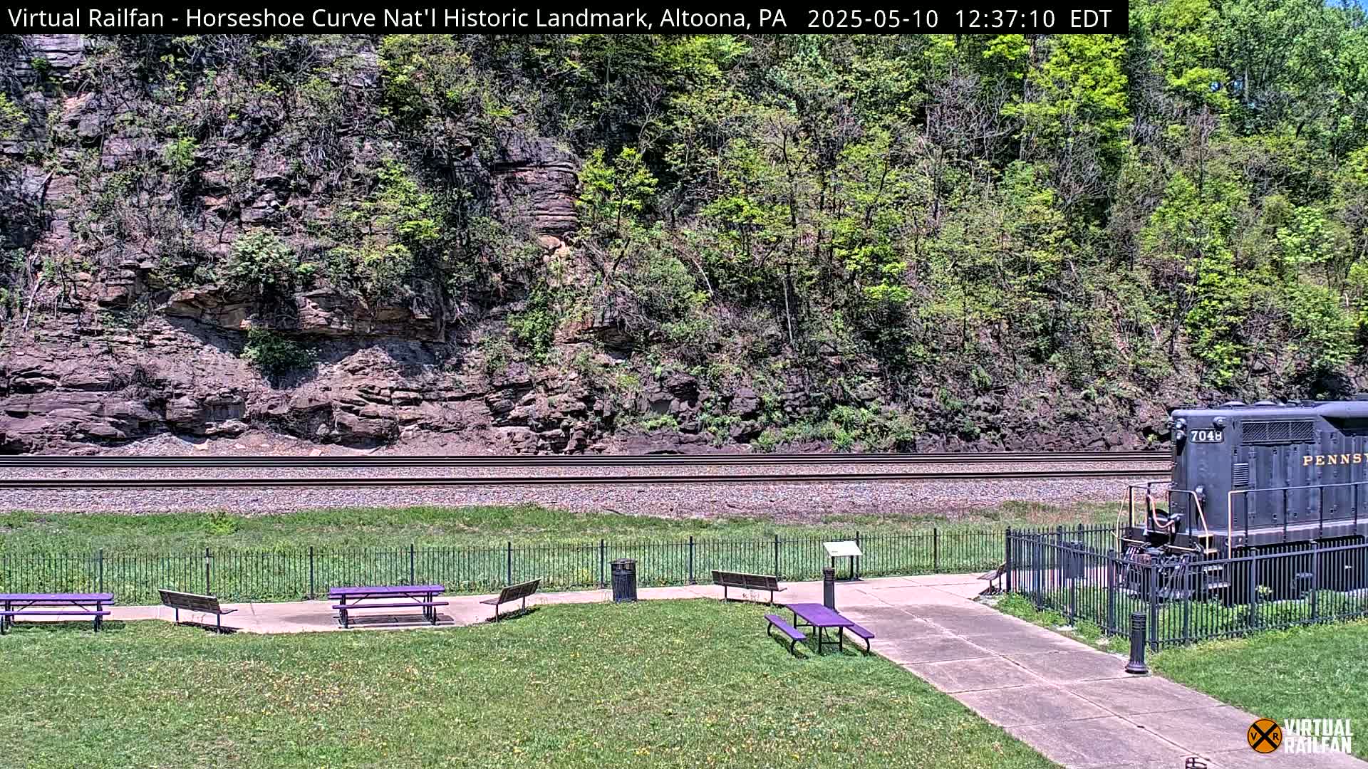 A railroad track runs in front of a rocky hillside covered in lush green vegetation, with a dark-colored locomotive partially visible at the right, picnic tables situated on a grassy area in the foreground on a sunny day.