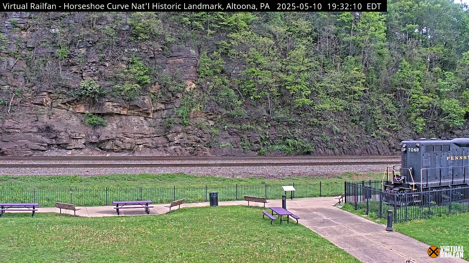 A partially visible train sits near a grassy area with picnic tables beside railroad tracks against a rocky hillside covered in lush green vegetation.