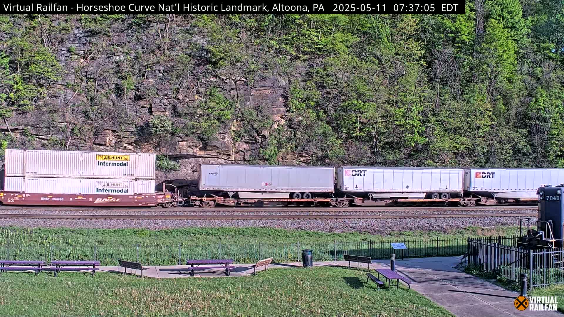 A freight train passes by a grassy area with picnic tables, against a backdrop of a rocky hillside covered in lush green trees under a sunny sky.