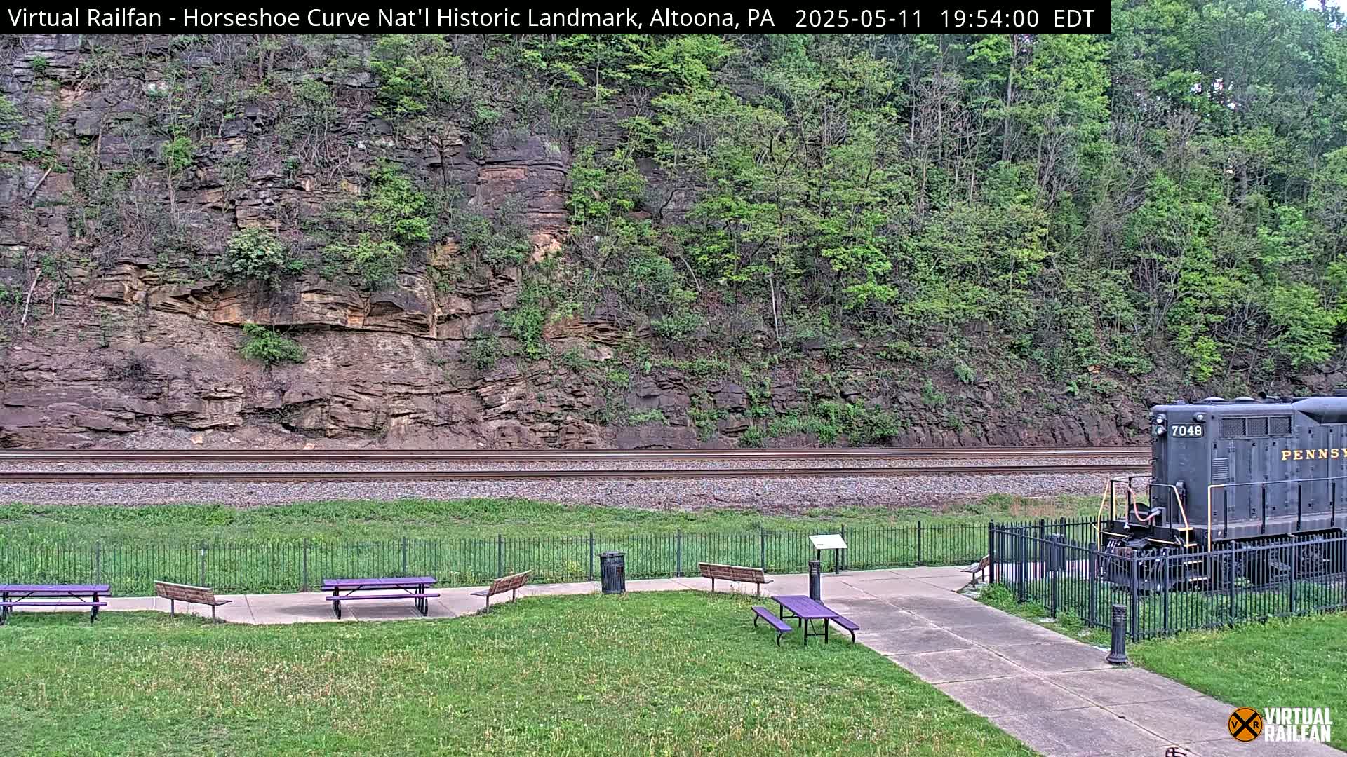 A dark-colored locomotive sits near a grassy area with picnic tables and a fence, beside railroad tracks running along a rocky hillside with lush green foliage under a daytime sky.