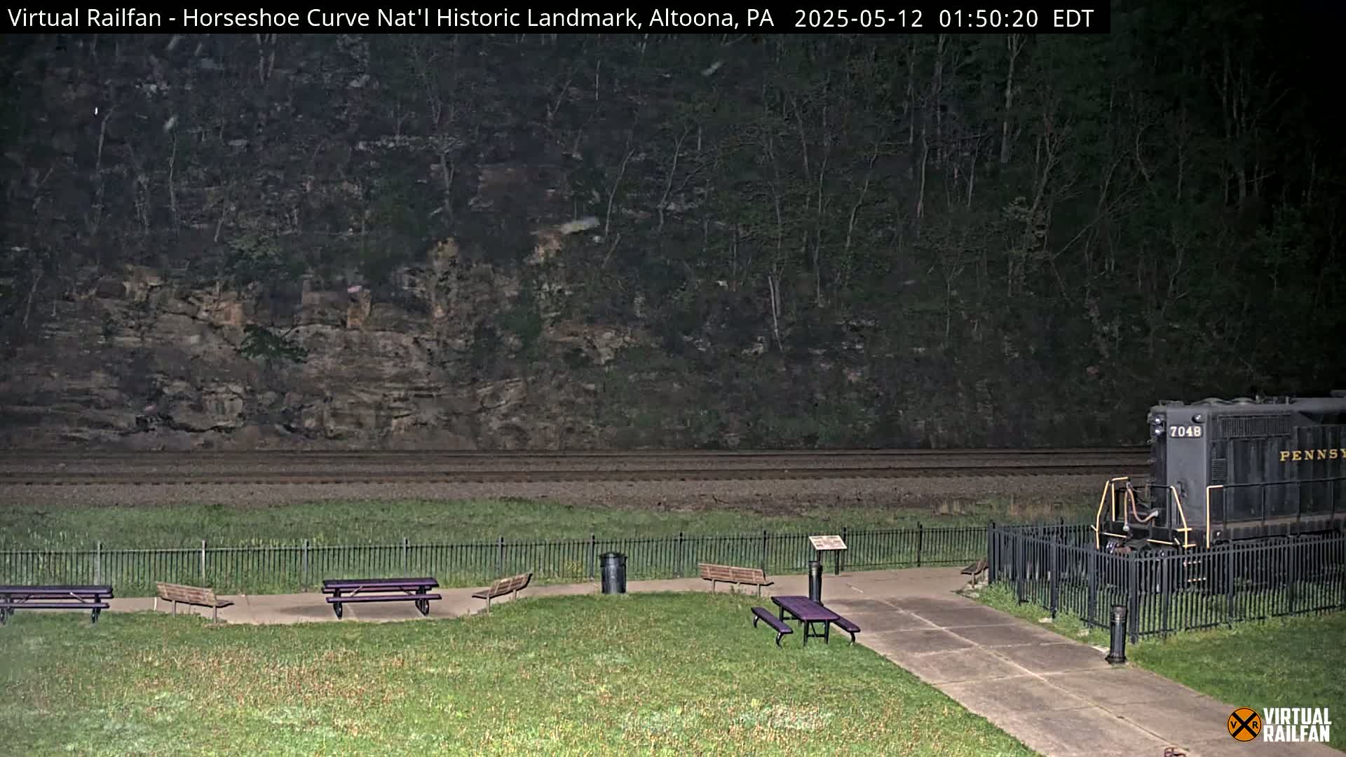 Under a dark night sky, a train sits behind a fence near a grassy area with several picnic tables.