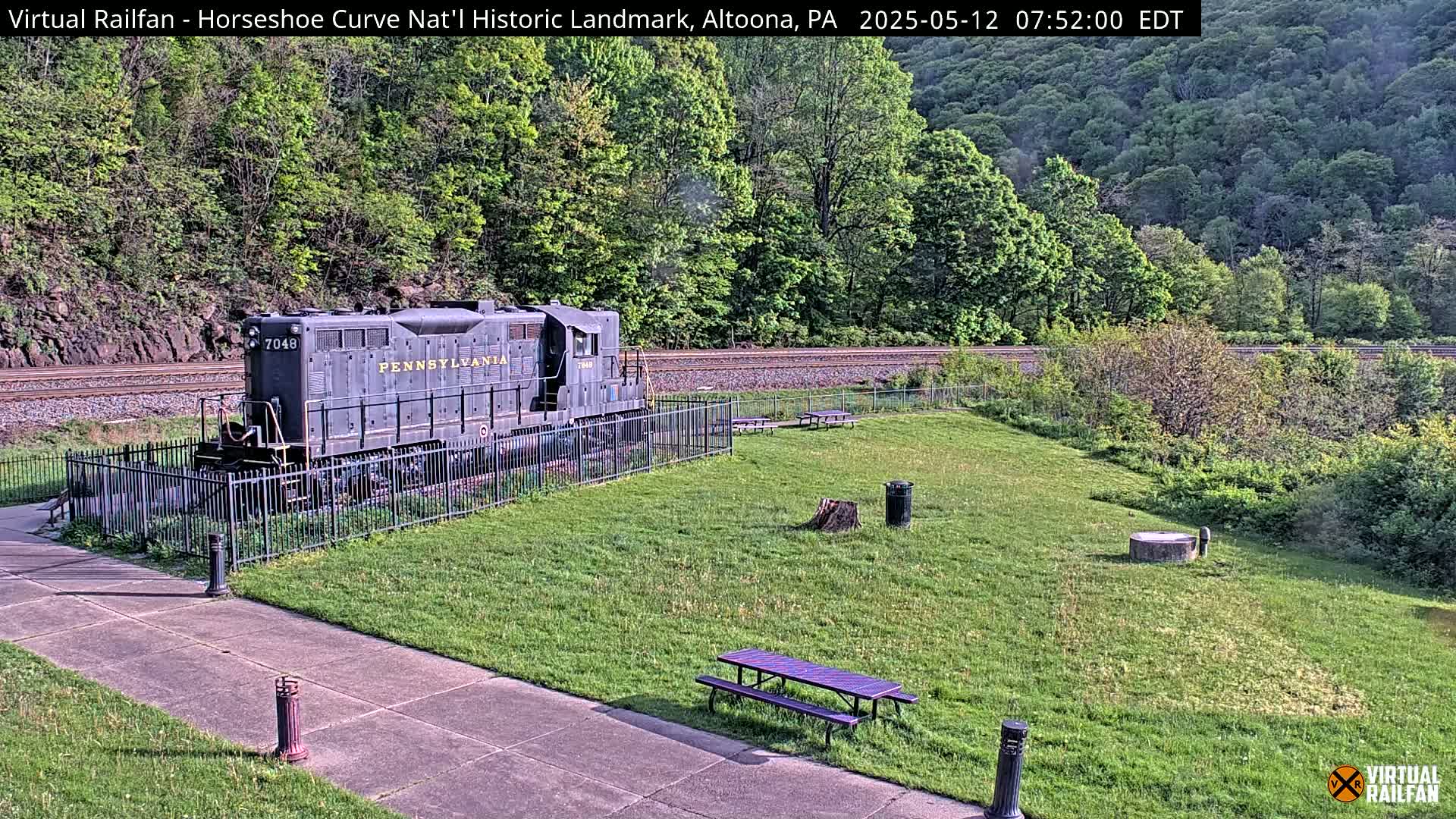 A dark-grey Pennsylvania Railroad locomotive sits behind a black metal fence on a grassy area with picnic tables, a paved walkway, and a background of lush green trees on a sunny day.