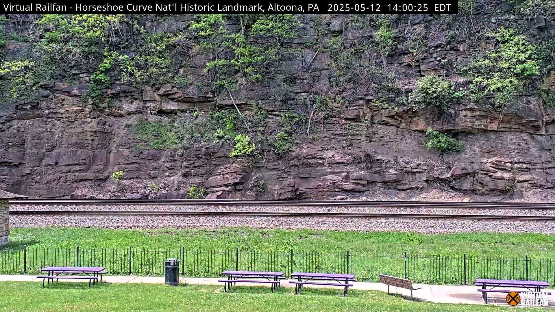 A rocky hillside with vegetation is visible behind a set of train tracks, and in the foreground, several purple picnic tables sit on a grassy area separated from the tracks by a fence.