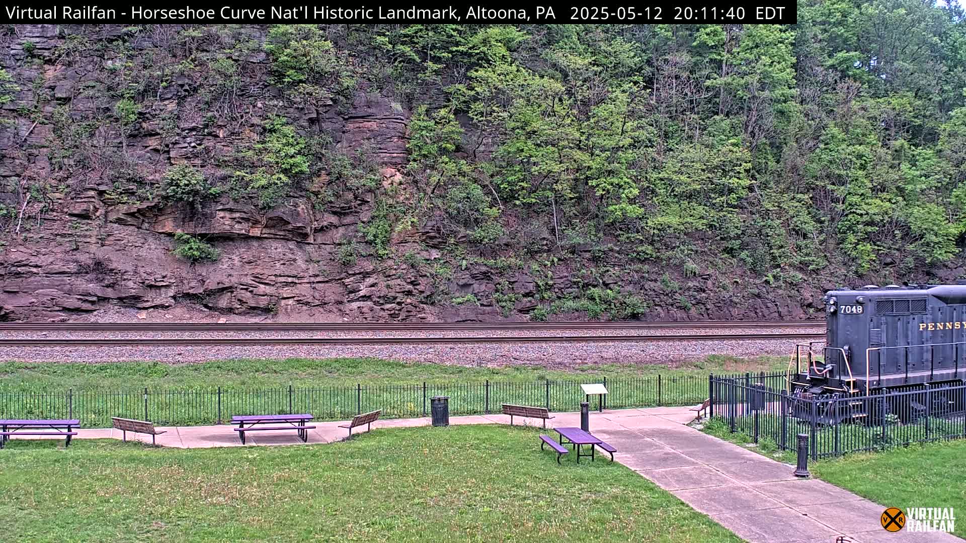 A train sits near a grassy area with picnic tables beside a railroad track that runs along the base of a rocky hillside covered in green trees and shrubs, under a daytime sky.
