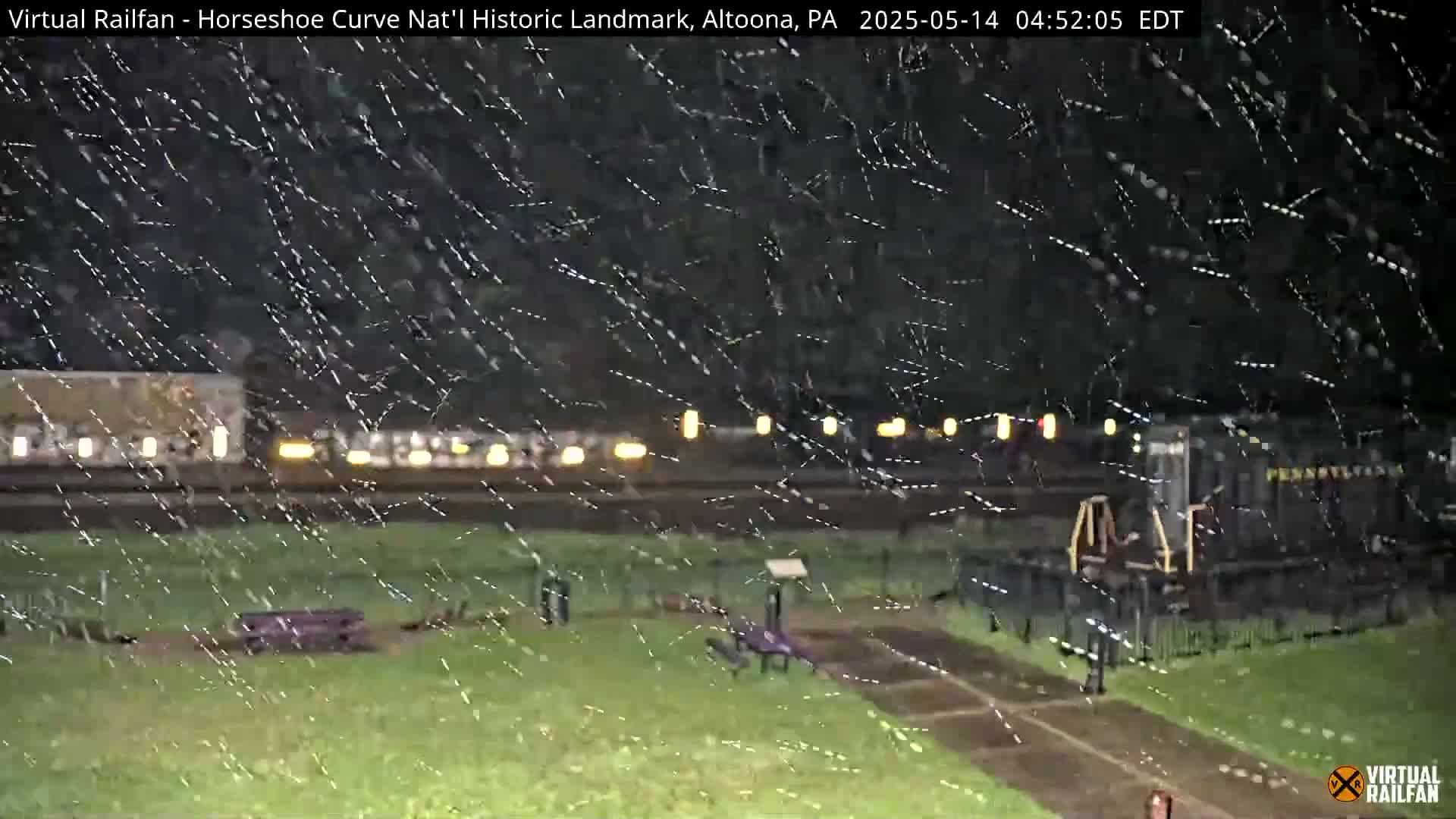 A nighttime scene of a train in the rain, viewed from across a grassy area.