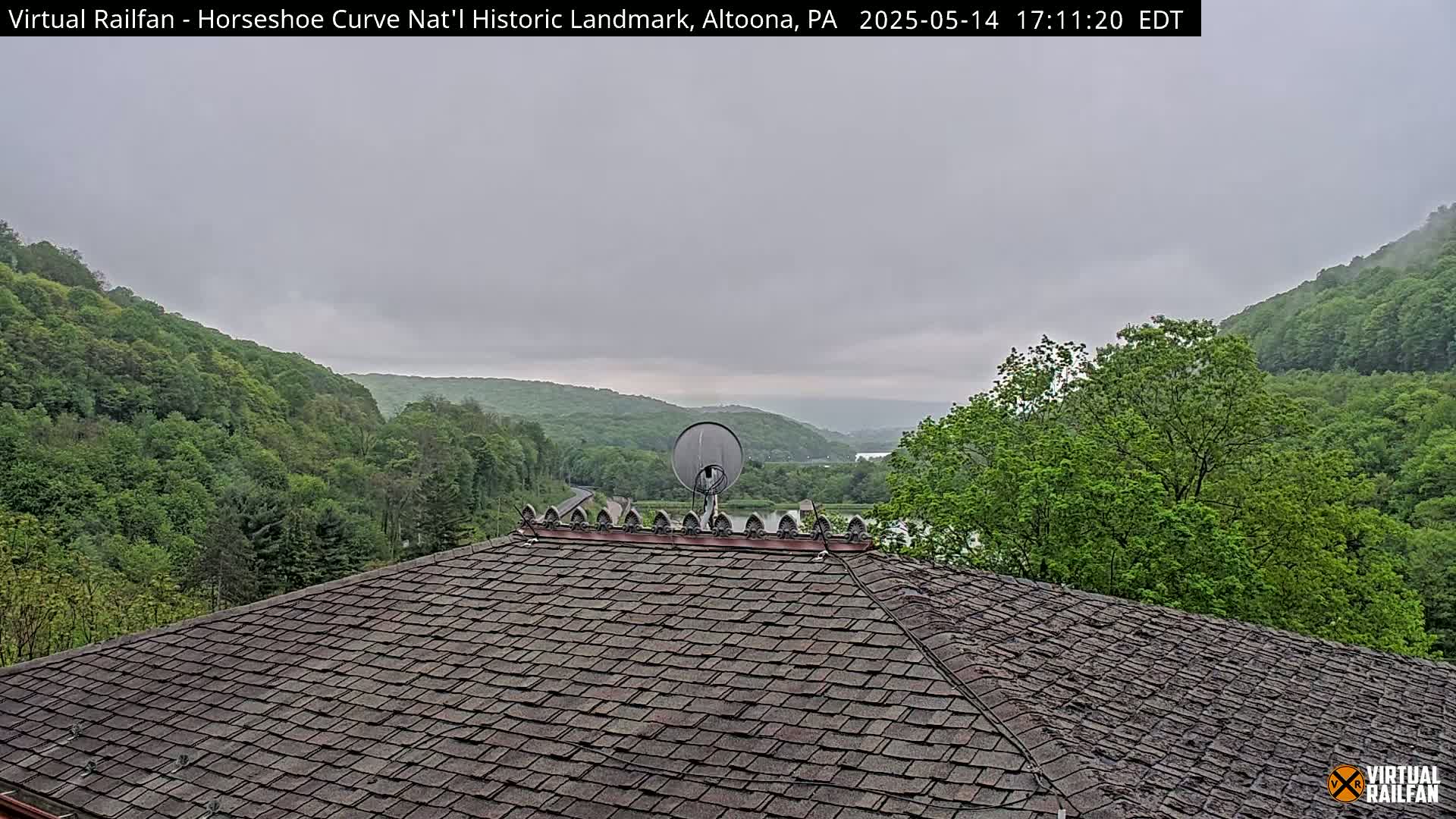 A dark gray, overcast sky hangs over a view of lush green hills and a valley, seen from a dark gray shingled roof with a satellite dish on top.