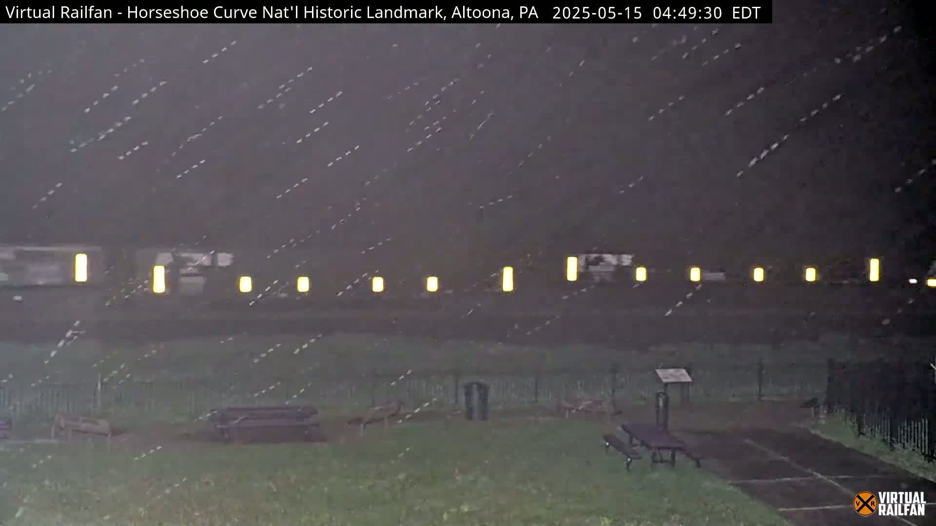 A nighttime scene of a park with picnic tables, under a heavy rain, shows illuminated markers along a distant track.