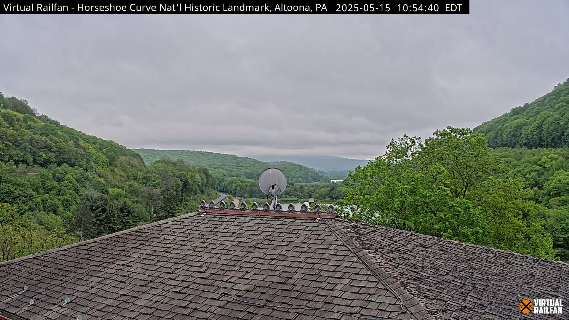 A dark gray overcast sky over lush green hills and a valley, viewed from a dark shingled roof with a satellite dish.