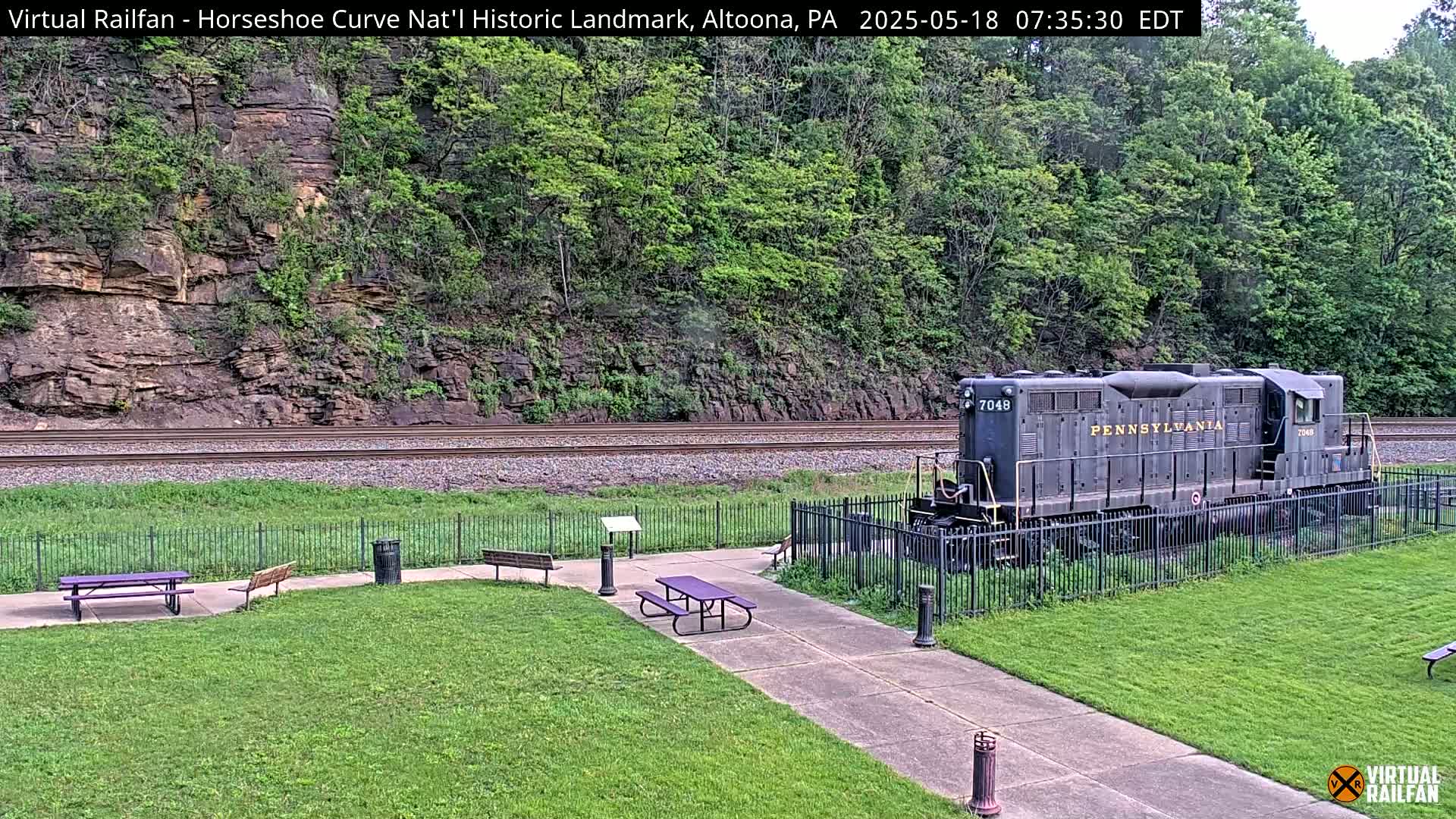 A Pennsylvania Railroad diesel locomotive sits behind a fence in a grassy park area next to railroad tracks and a steep, tree-covered hillside on a partly sunny day.